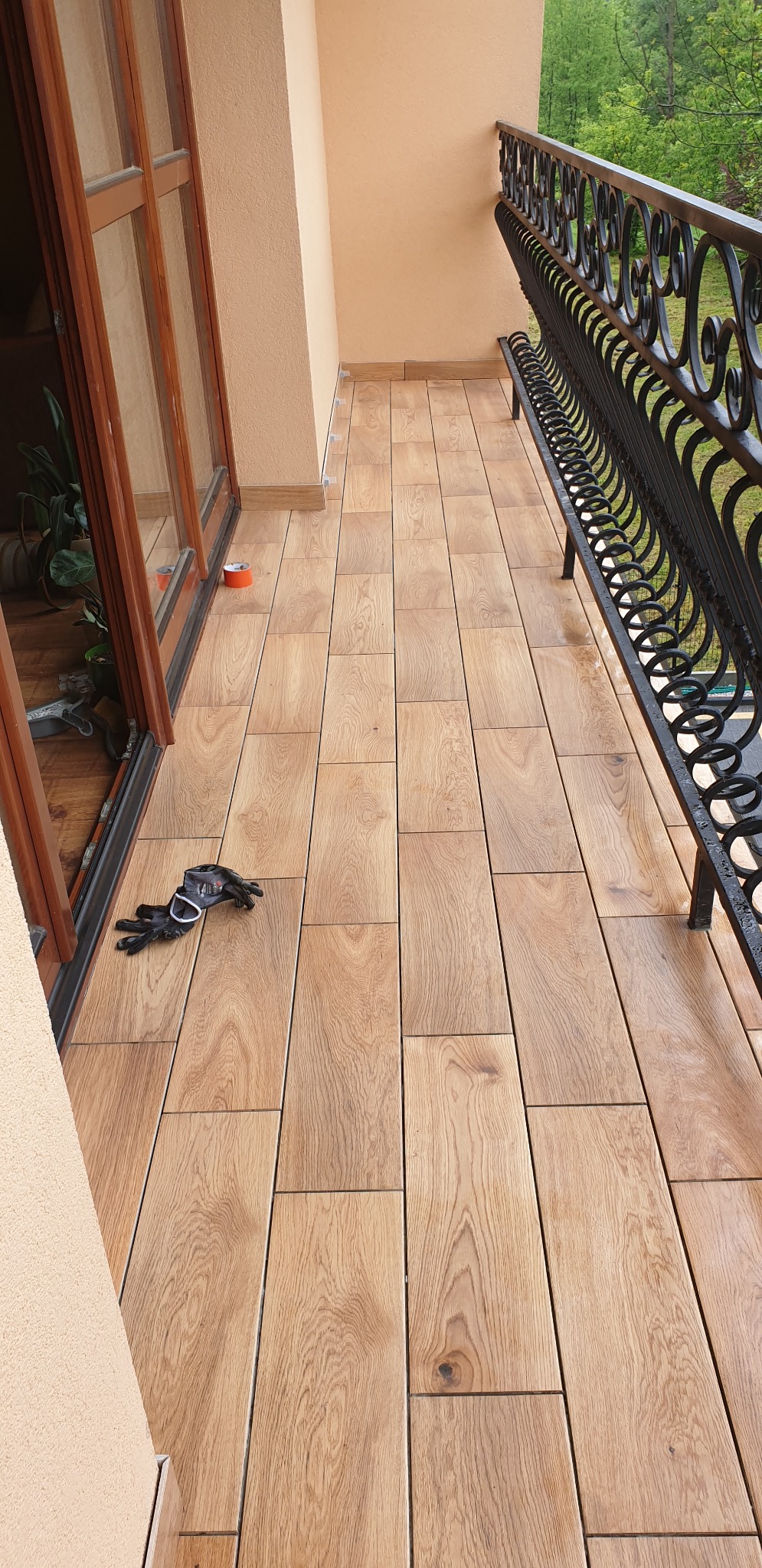 Ułożona podłoga z drewnopodobnych płytek na balkonie, widoczne czarne rękawice robocze, taśma malarska i ozdobna, metalowa balustrada.