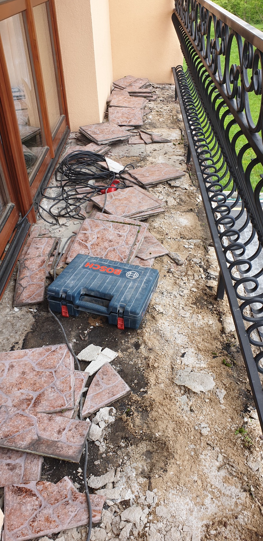 Demontaż brązowych płytek na balkonie, widoczna skrzynka narzędziowa Bosch, przewody elektryczne i fragment ozdobnej balustrady.