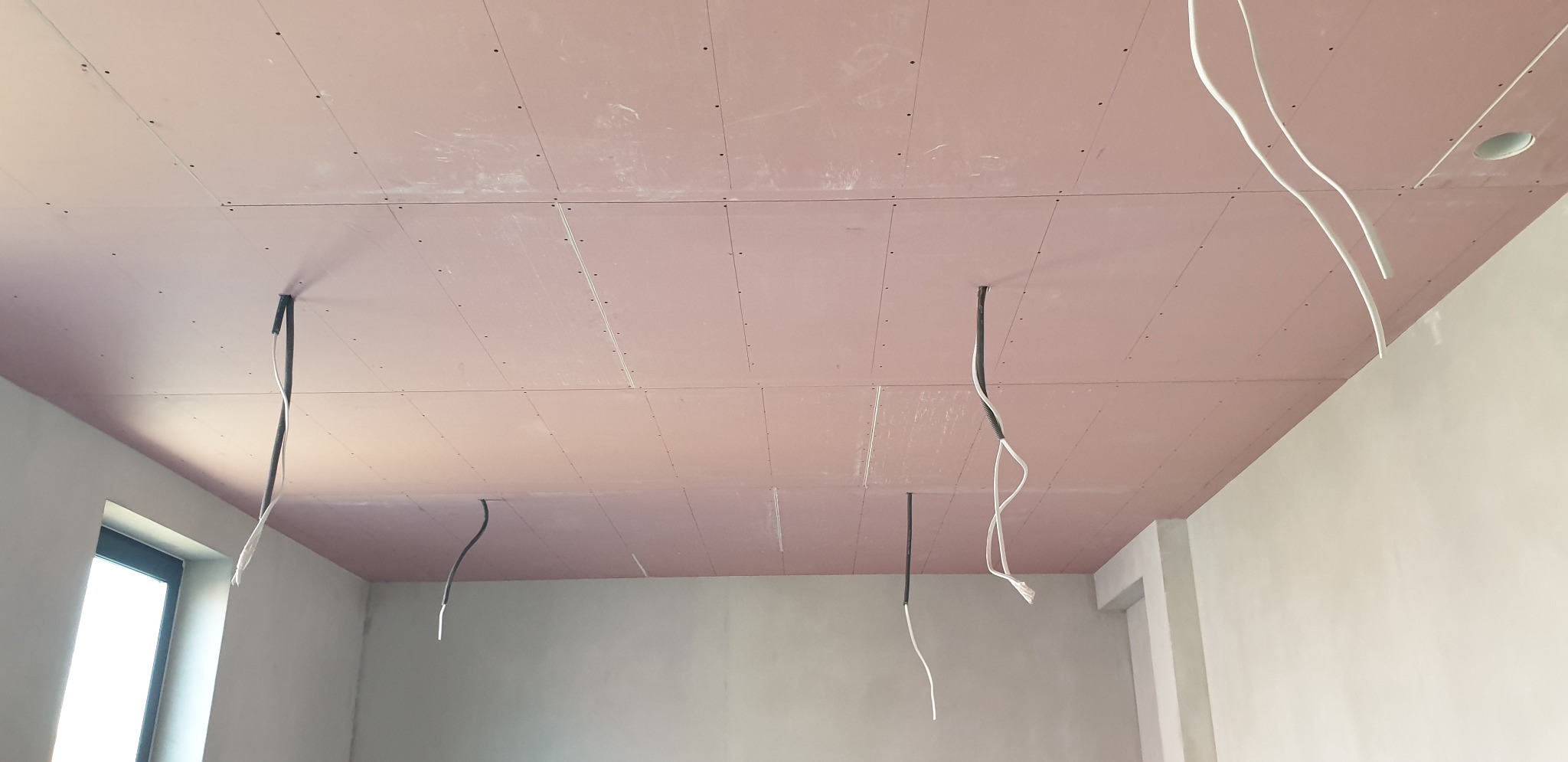 Różowa płyta gipsowo-kartonowa na suficie z wystającymi przewodami elektrycznymi w surowym wnętrzu.
