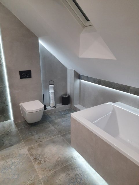 Nowoczesna łazienka na poddaszu z podświetleniem LED, wiszącą toaletą i wanną obudowaną płytkami w odcieniach szarości oraz wzorzystą podłogą.