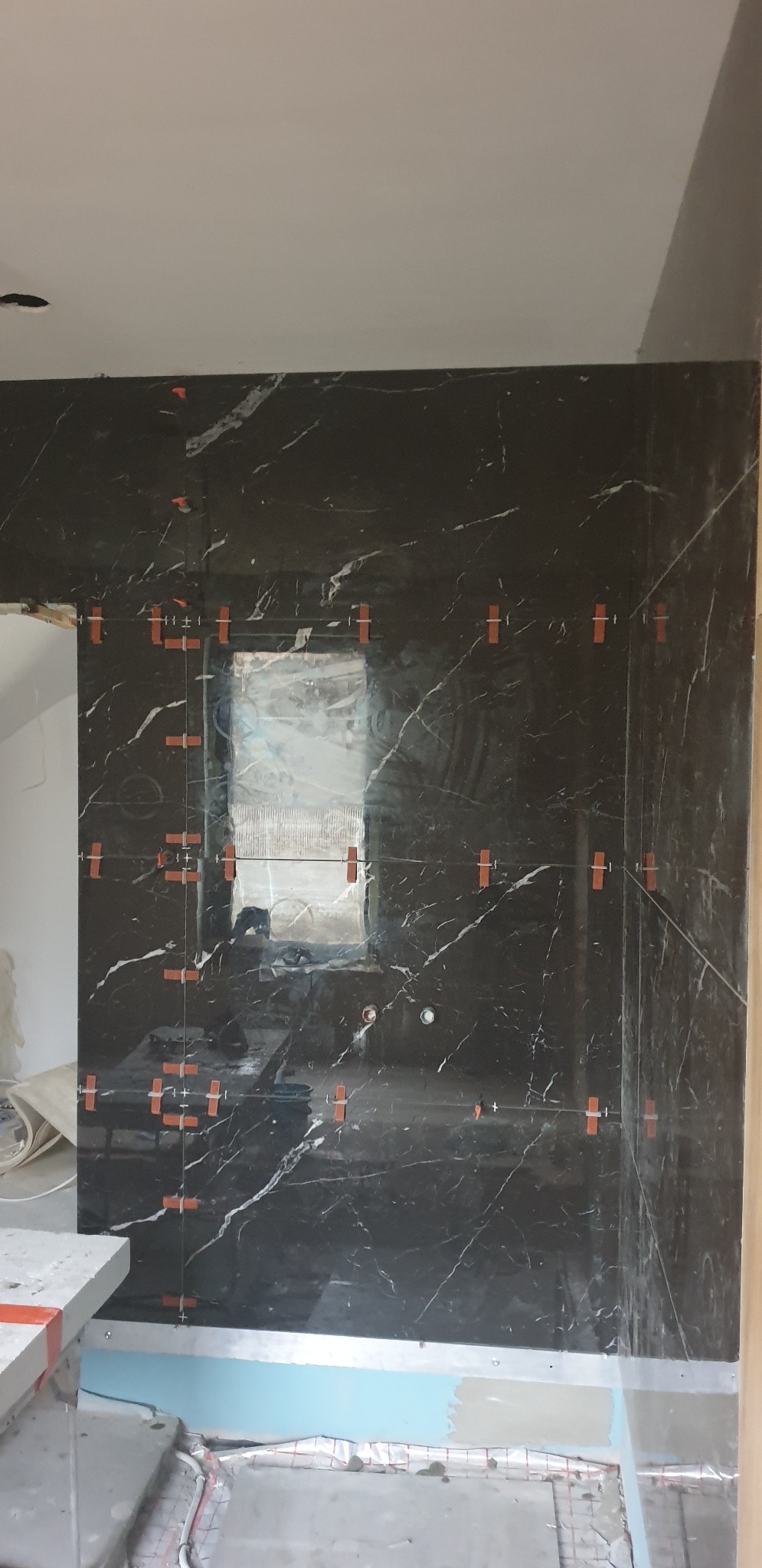 Częściowy widok ściany łazienki z czarnych płytek imitujących marmur, z pomarańczowymi krzyżykami dystansowymi, w trakcie procesu układania, z widocznym ogrzewaniem podłogowym.