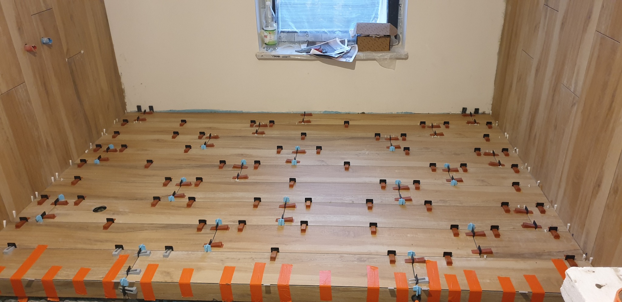 Układanie płytek imitujących drewno na podłodze w trakcie prac wykończeniowych, z zastosowaniem klipsów poziomujących i taśmy maskującej przy ścianie.