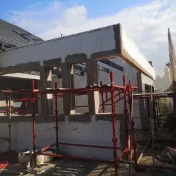 Budynek w trakcie ocieplania ścian zewnętrznych, widoczne szare płyty izolacyjne i czerwone rusztowania, etap prac przygotowawczych.