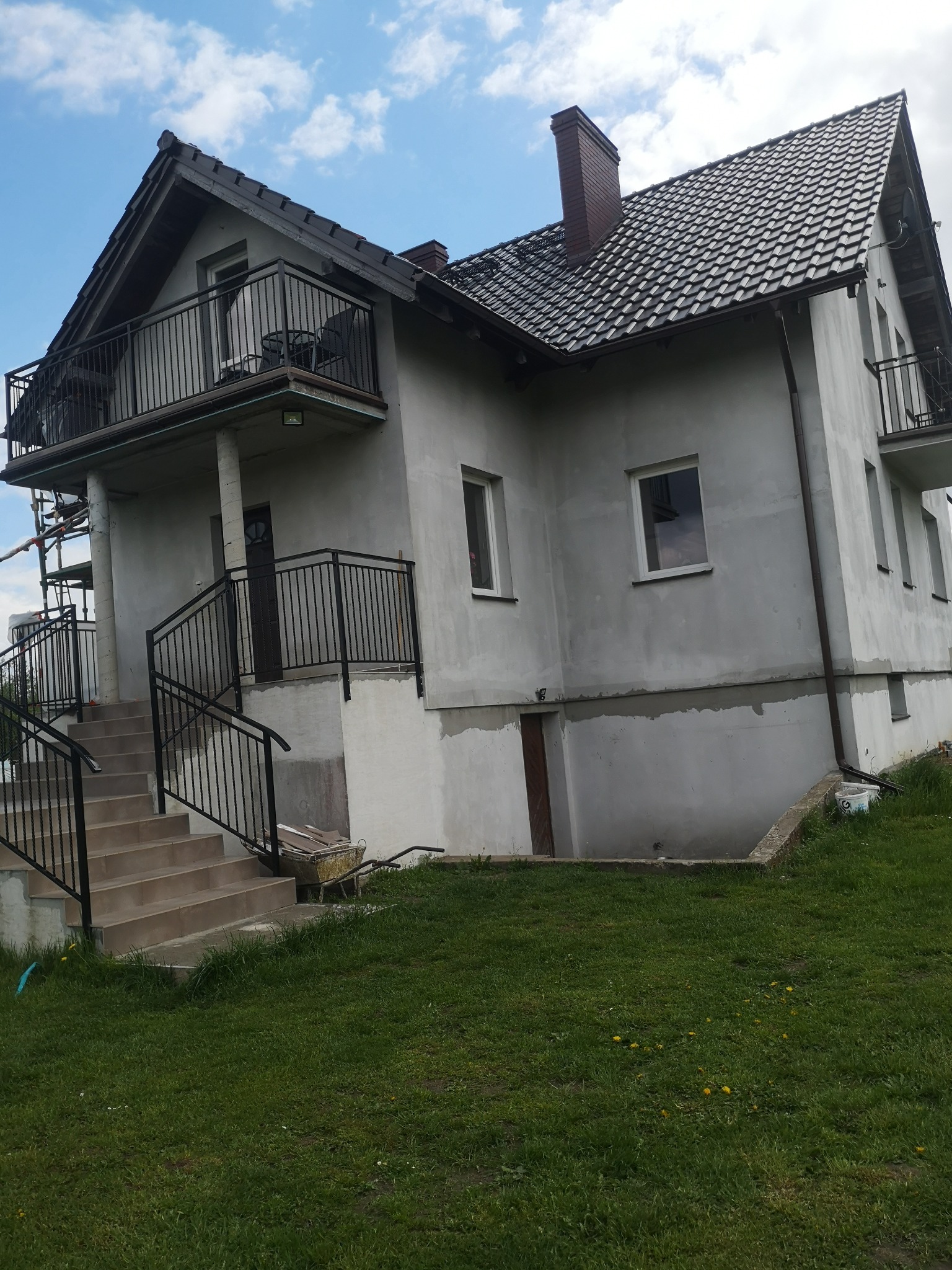 Dom jednorodzinny z szarą elewacją w trakcie prac wykończeniowych, widoczne rusztowania i taczka z zaprawą, trawnik z mleczami.