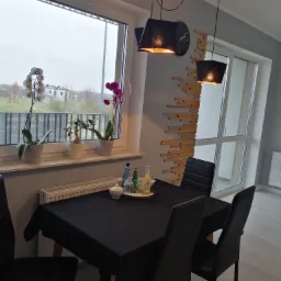 Nowoczesne wnętrze jadalni z czarnym stołem nakrytym czarnym obrusem, otoczonym czarnymi krzesłami, z oknem z widokiem na zewnątrz, dekoracjami z drewna na ścianie i wiszącymi lampami.