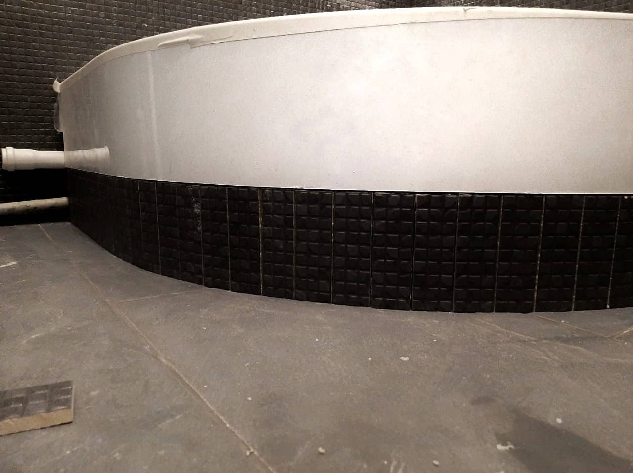 Niski kąt widzenia na obudowę wanny z czarnych, kwadratowych płytek mozaikowych, biała góra obudowy zabezpieczona taśmą malarską, szara podłoga i fragment czarnej ściany w tle.