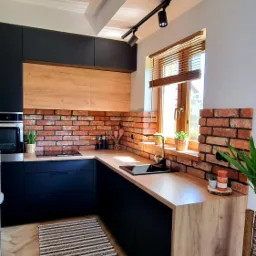 Nowoczesna kuchnia z czarnymi szafkami, blatem z drewna i ceglaną ścianą, oświetlona reflektorami punktowymi, z dywanikiem w paski na parkiecie ułożonym w jodełkę.