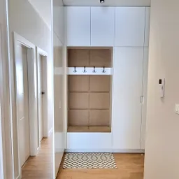 Nowoczesny przedpokój z białymi szafami w zabudowie, wnęką z siedziskiem obitym beżowym materiałem, wieszakami i dywanikiem w geometryczny wzór na podłodze z paneli.