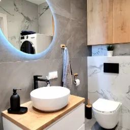 Nowoczesna łazienka z okrągłym lustrem podświetlanym LED, umywalką nablatową na szafce z drewnianym blatem, wiszącą toaletą i szarą płytką imitującą marmur na ścianach.