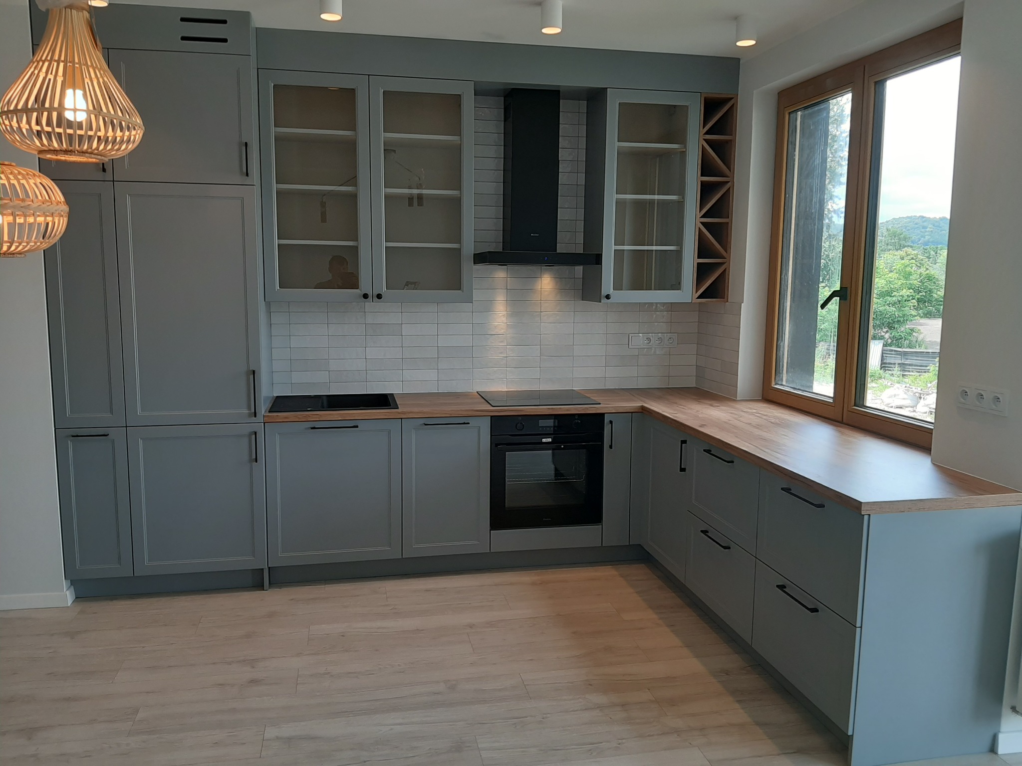 Nowoczesna kuchnia w odcieniach szarości z drewnianym blatem, oświetlona wiszącymi lampami z naturalnego materiału. Widok na zielony krajobraz za oknem.