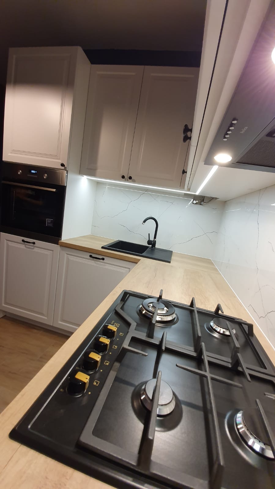 Nowoczesna kuchnia z białymi szafkami, czarnym zlewem i płytą gazową z żółtymi detalami na tle marmurowej ściany.