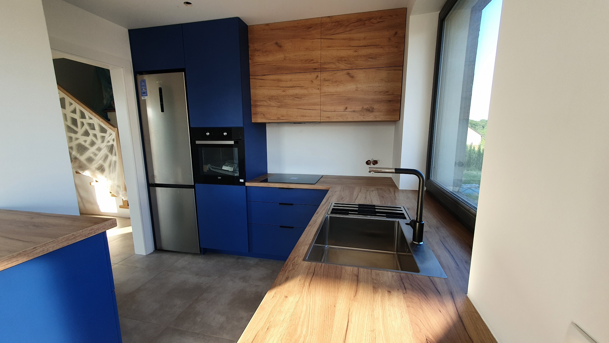 Nowoczesna kuchnia: granatowe szafki, drewnopodobne blaty, srebrna lodówka i piekarnik, widok na schody zabezpieczone folią.