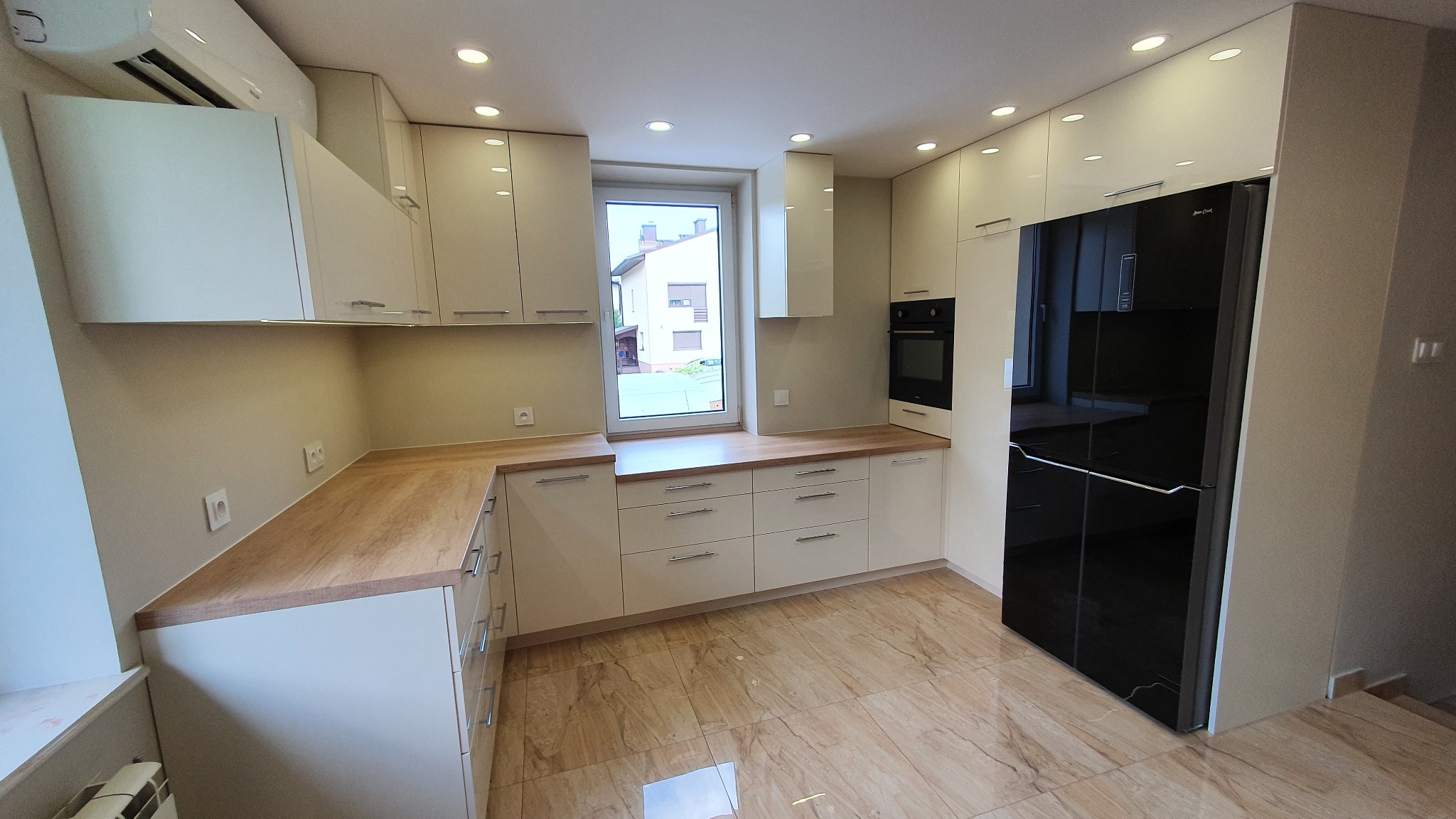 Jasna kuchnia na wymiar z drewnianym blatem, czarną lodówką side-by-side i oknem z widokiem na budynek mieszkalny.