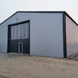 Nowoczesna hala magazynowa z szarej blachy trapezowej z czarnymi detalami wykończeniowymi, widoczne drzwi wejściowe i brama wjazdowa, teren wokół hali częściowo utwardzony, ślady opon na piasku.