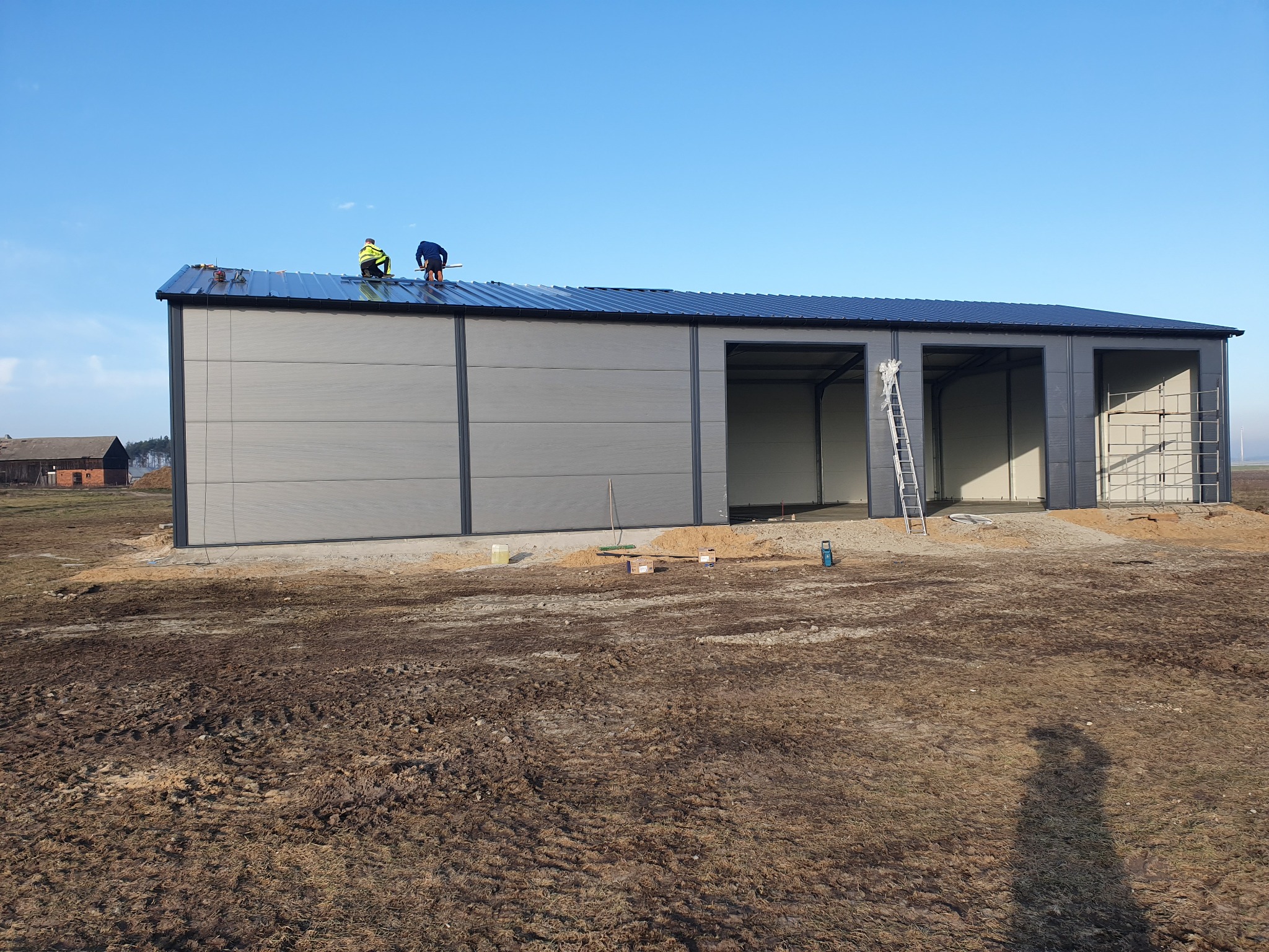 Montaż hali z płyt warstwowych: dwaj pracownicy na dachu, widoczne panele ścienne i konstrukcja stalowa, teren budowy z błotem.