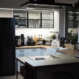 Nowoczesna kuchnia z białą zabudową, drewnianym blatem, czarną lodówką i wyspą kuchenną z metalowym stelażem, oświetleniem LED pod szafkami i roślinami doniczkowymi.