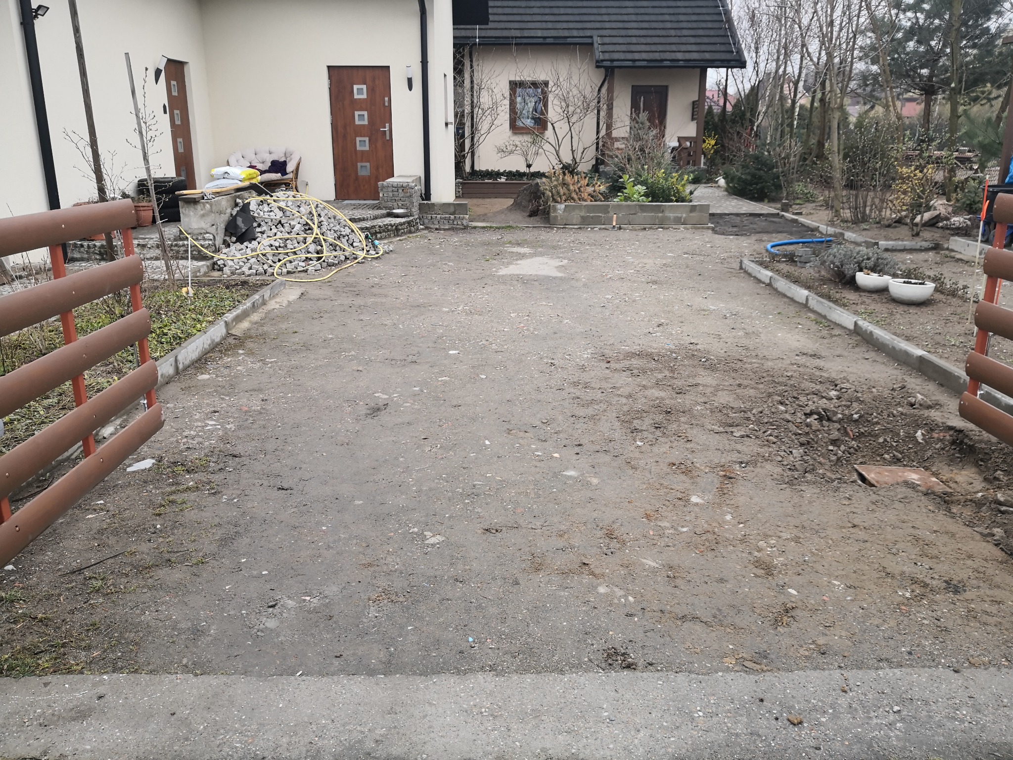 Przygotowanie terenu pod przyszły podjazd: widoczny stos kostki, rozłożony wąż, częściowo ułożone obrzeża i brakująca nawierzchnia