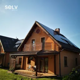 Dwa domy jednorodzinne z panelami słonecznymi na dachach, widoczne w słoneczny dzień, z logo firmy na górze kadru.