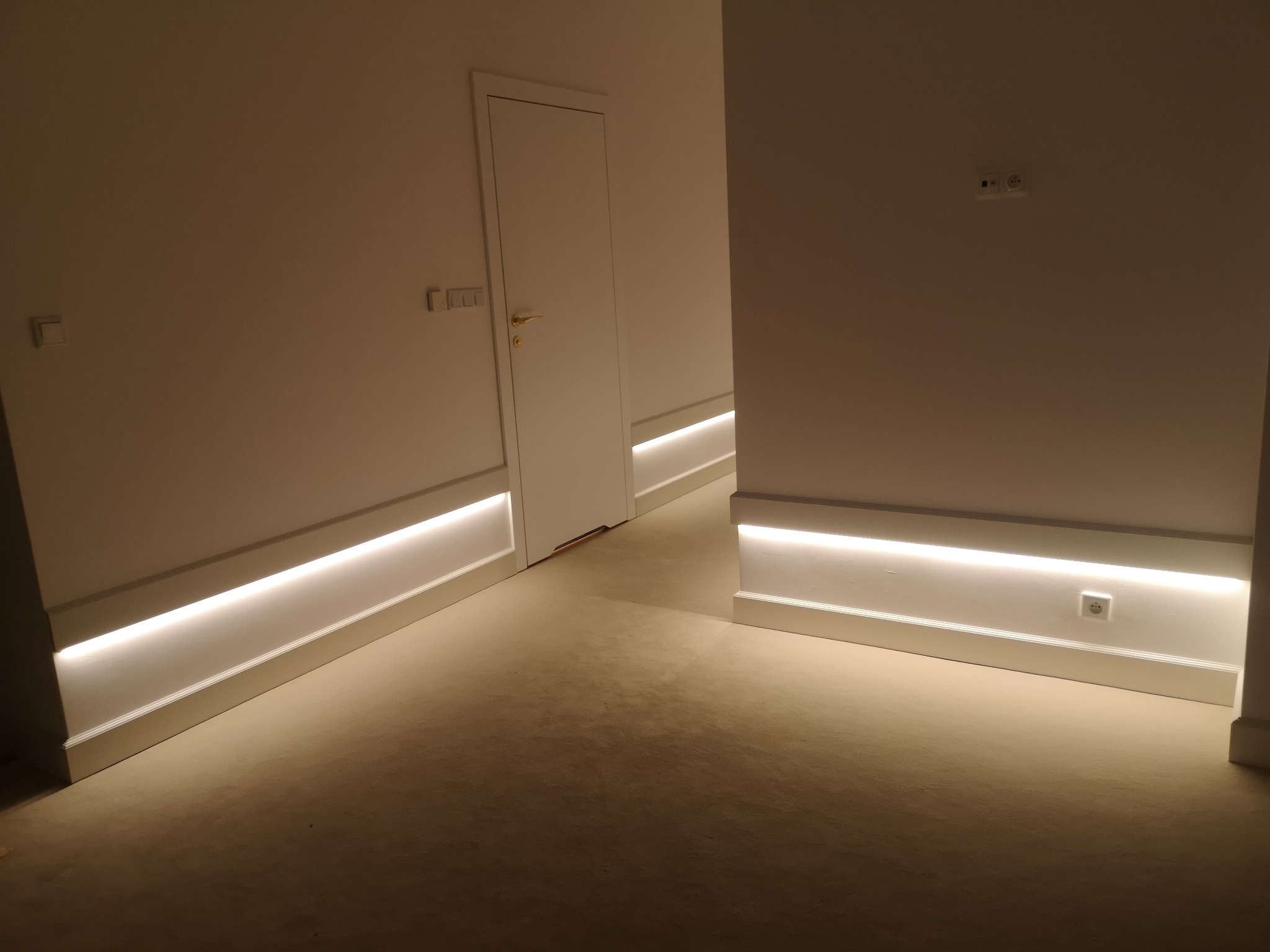 Nowoczesne oświetlenie przypodłogowe LED w korytarzu z białymi ścianami i drzwiami, widoczne gniazdka elektryczne i włączniki.