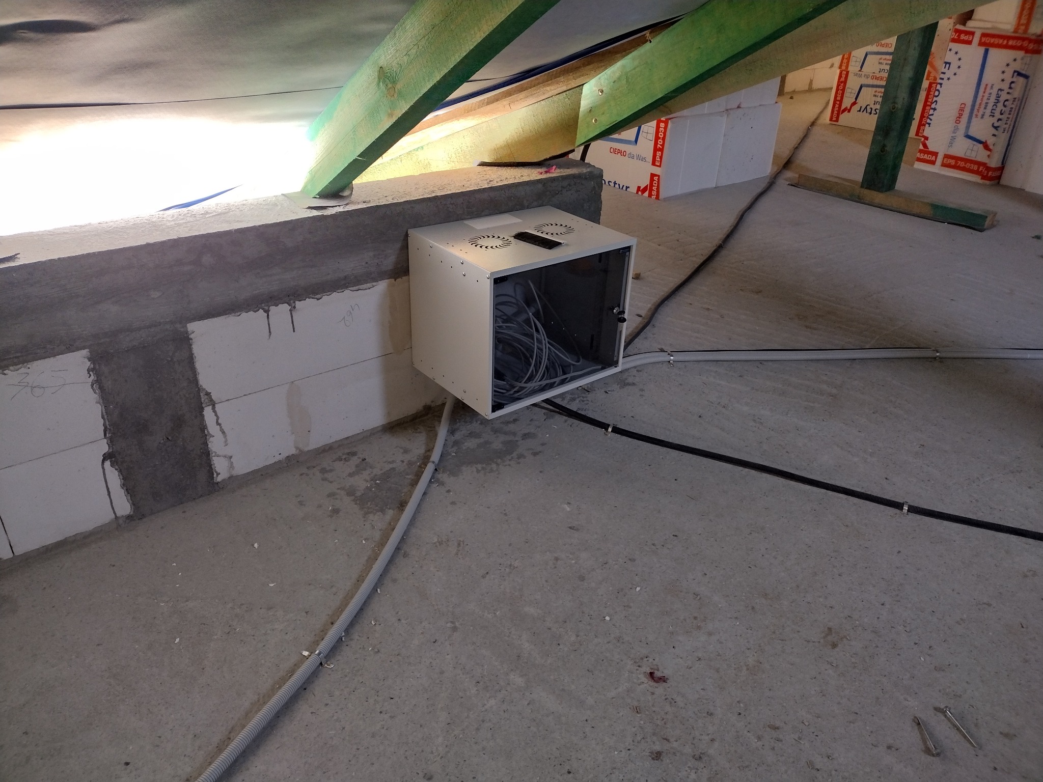 Biała szafka rack z widocznymi kablami wewnątrz, umieszczona w surowym pomieszczeniu z betonową podłogą i ścianami z pustaków, zielone elementy konstrukcyjne dachu w tle, widoczne przewody...