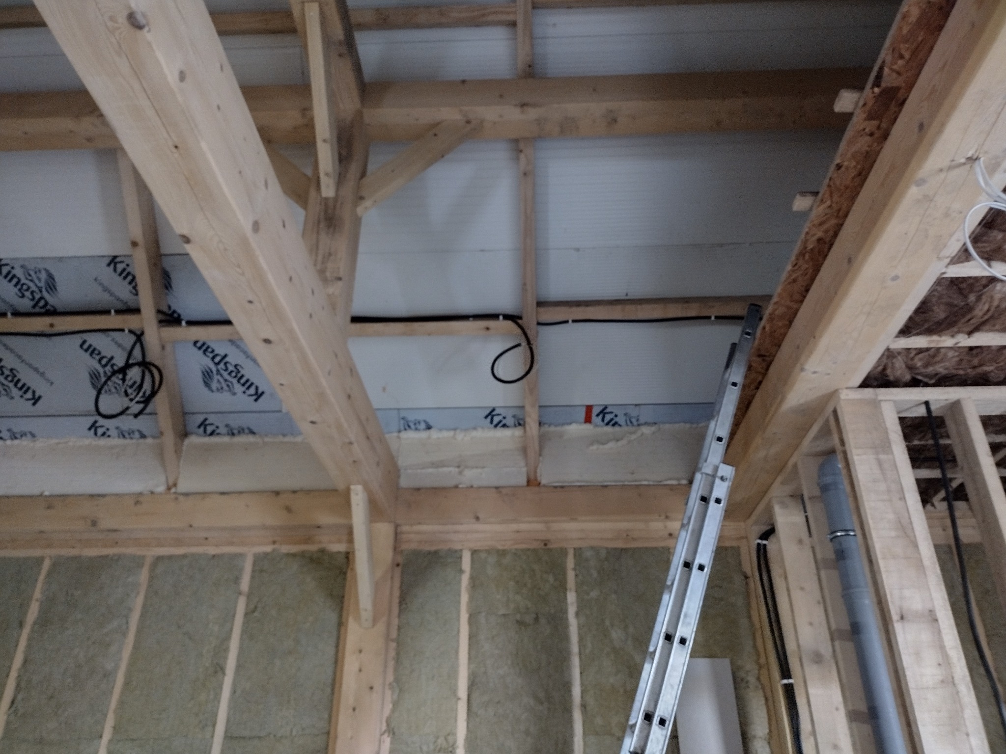 Wnętrze budynku w trakcie budowy: drewniana konstrukcja stropu z widocznymi przewodami elektrycznymi, izolacja z wełny mineralnej i aluminiowa drabina oparta o belkę.