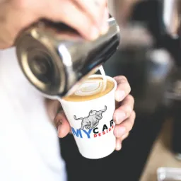 Barista wlewa spienione mleko do filiżanki z kawą, tworząc wzór latte art. Na filiżance widoczne logo 'MYCAR DESIGN' z motywem byka.