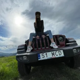 Kobieta siedzi na masce bordowego Jeepa z tablicą rejestracyjną S1 MCD na tle zachmurzonego nieba i zielonej trawy