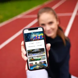 Kobieta na bieżni trzyma telefon z otwartą stroną mycardesign.pl, prezentującą zdjęcia i filmy samochodów oraz motocykli.