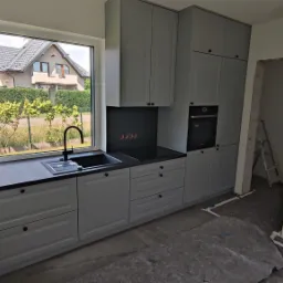 Szara zabudowa kuchenna z czarnym blatem i zlewozmywakiem pod oknem z widokiem na dom i zieleń, widoczna płyta indukcyjna i piekarnik, podłoga zabezpieczona folią malarską.