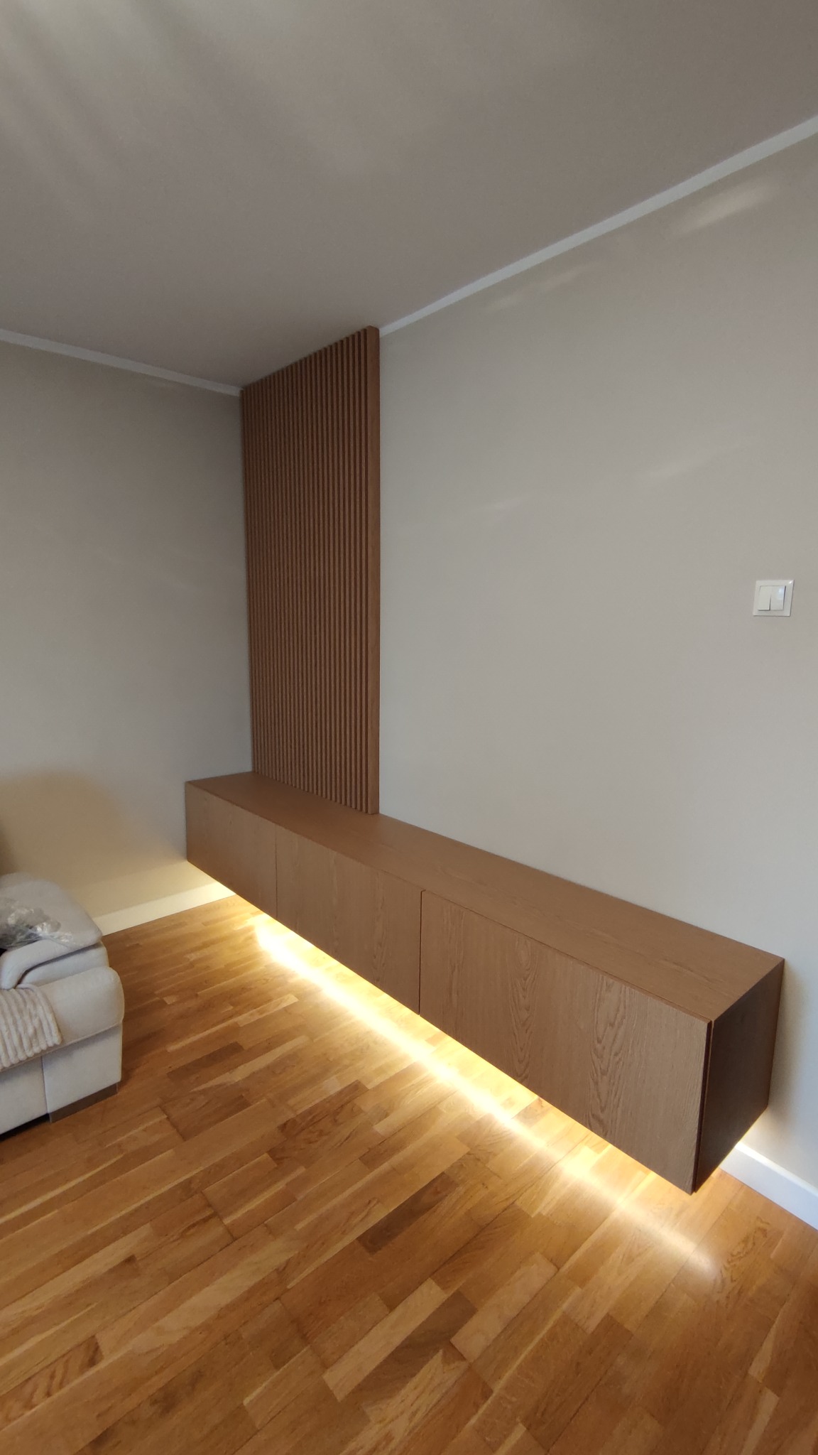 Nowoczesna szafka RTV z podświetleniem LED pod spodem, drewniane lamele na ścianie, parkiet w jodełkę.