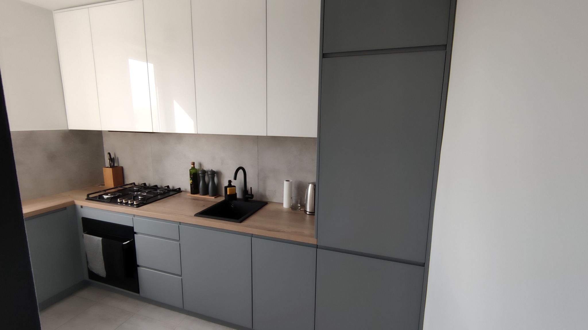 Nowoczesna kuchnia z szarymi dolnymi szafkami i białymi górnymi, czarny zlew pod oknem, drewniany blat, płyta gazowa i szara ściana nad blatem.