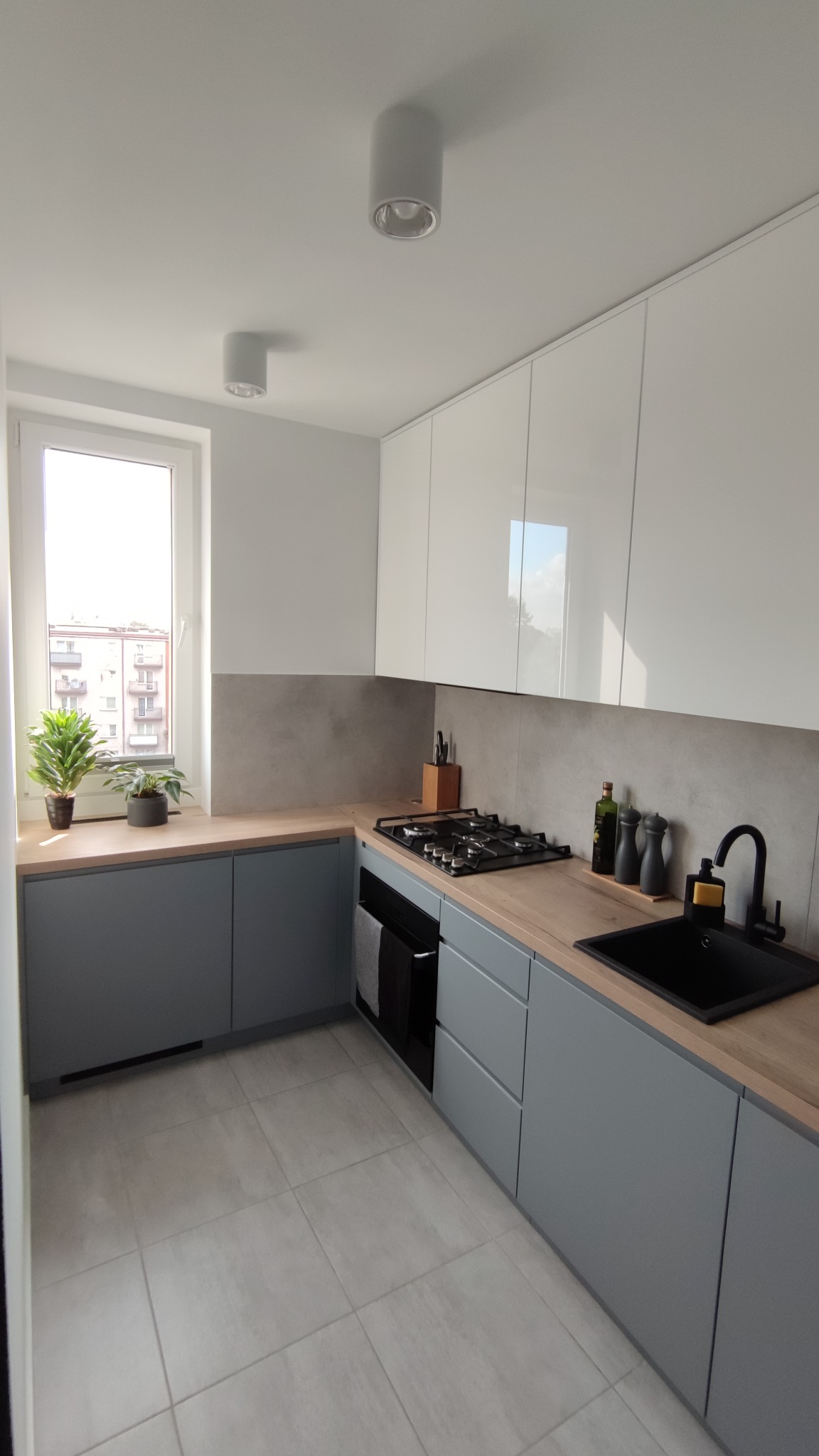 Nowoczesna, jasna kuchnia z białymi szafkami górnymi i szarymi dolnymi, drewnianym blatem, czarnym zlewem i armaturą, z widokiem na budynek mieszkalny za oknem.
