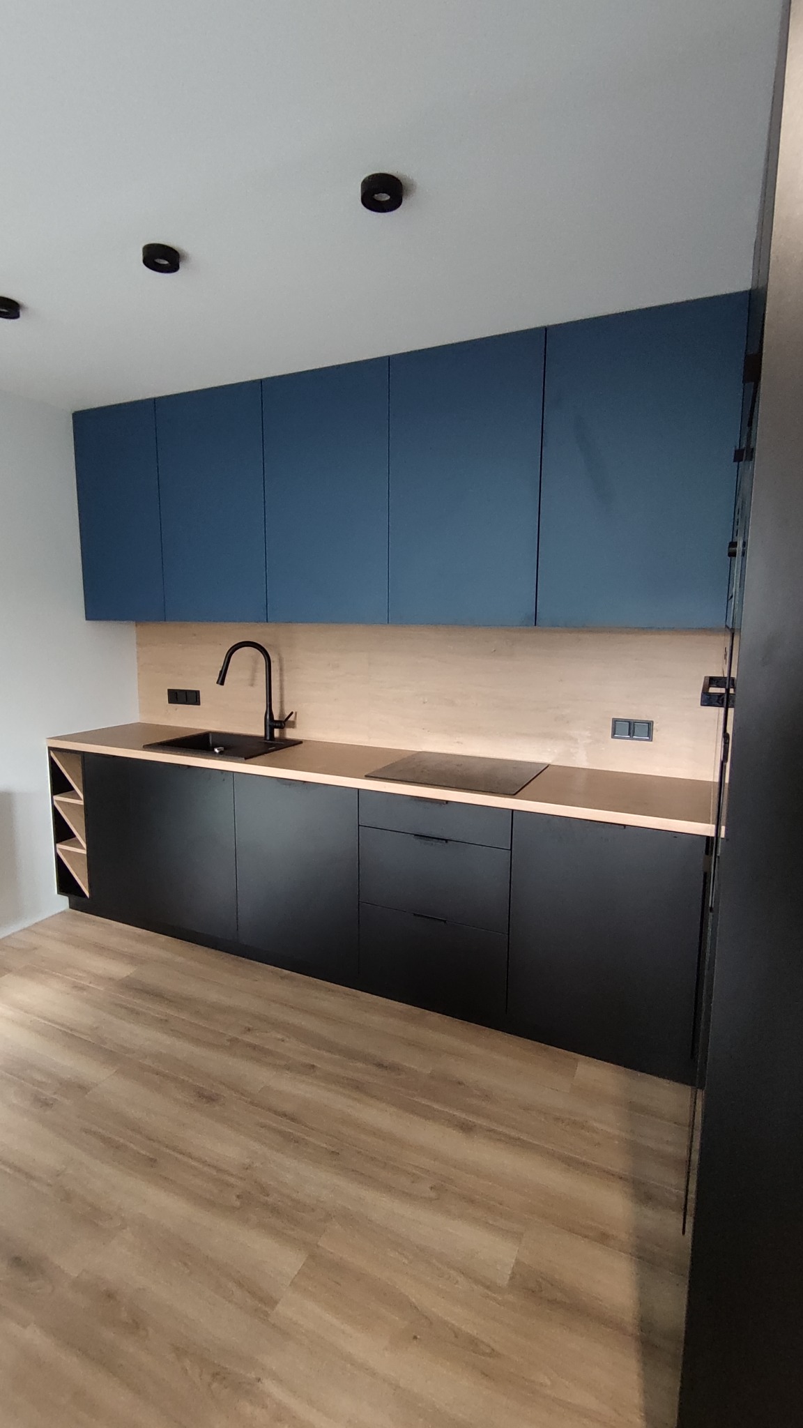 Nowoczesna kuchnia z ciemnymi szafkami dolnymi i niebieskimi górnymi, drewnianym blatem i panelem ściennym, czarną armaturą i płytą indukcyjną.