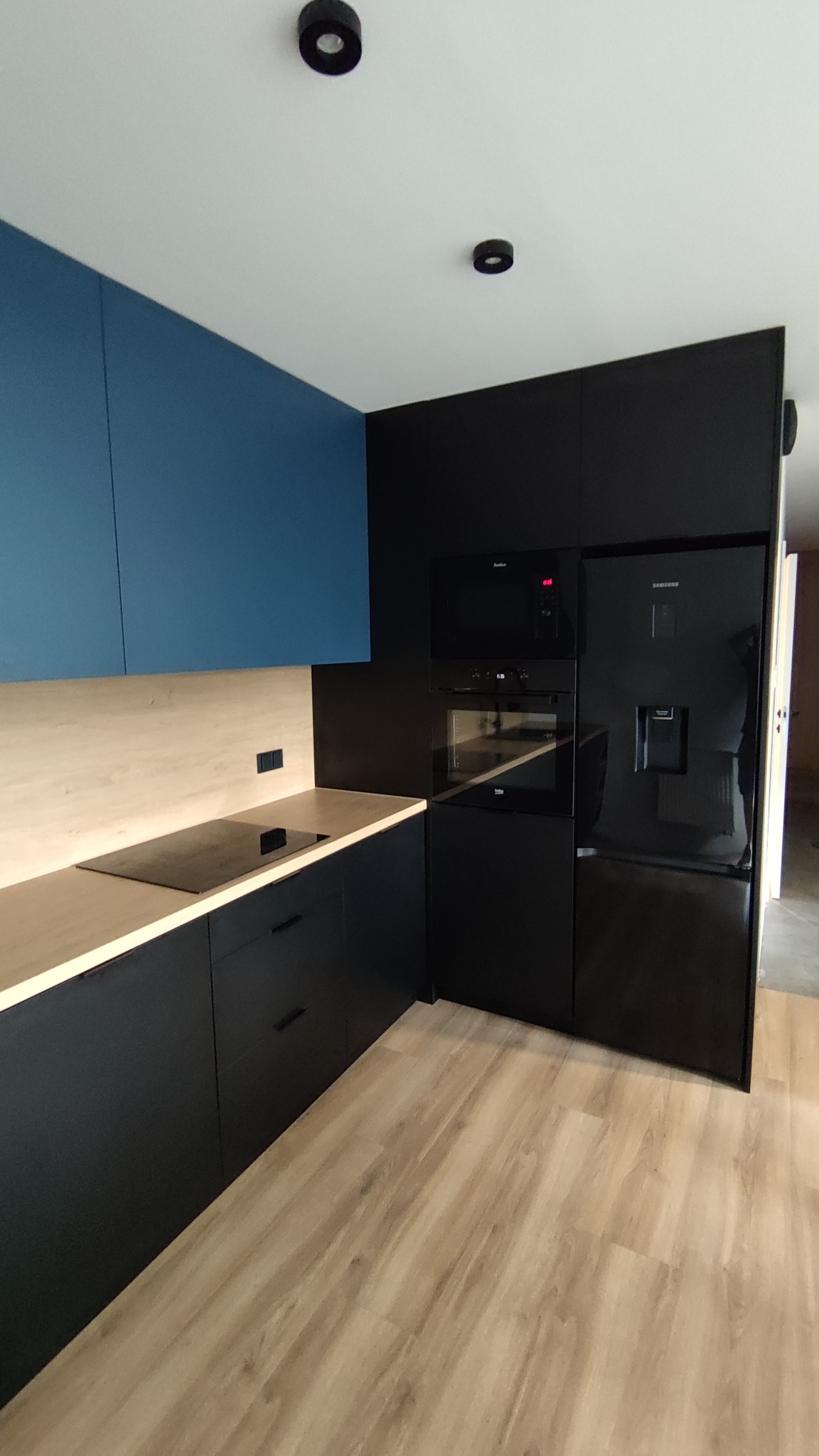 Nowoczesna kuchnia z niebieskimi szafkami górnymi, drewnianym blatem, czarnymi szafkami dolnymi i sprzętem AGD w zabudowie.