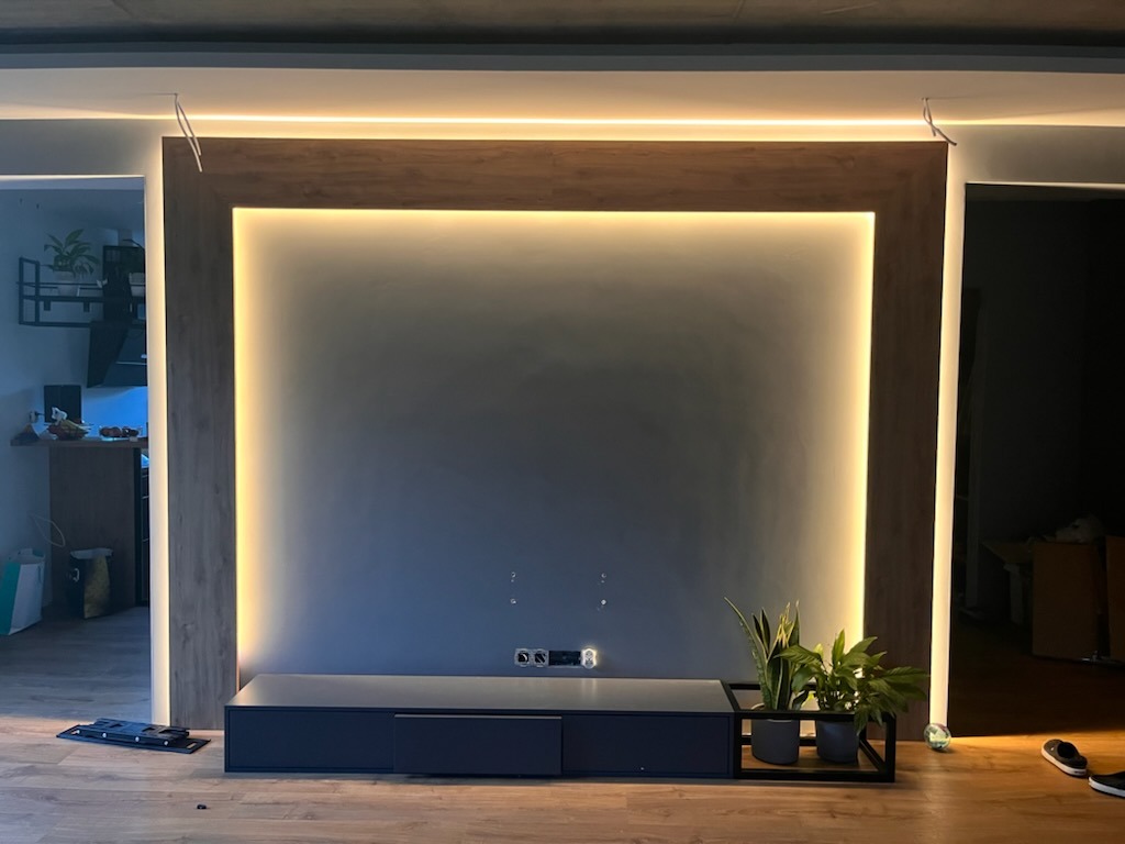 Panel ścienny z drewna z podświetleniem LED, pod nim czarna szafka RTV i rośliny doniczkowe, widoczne gniazdka elektryczne na ścianie.