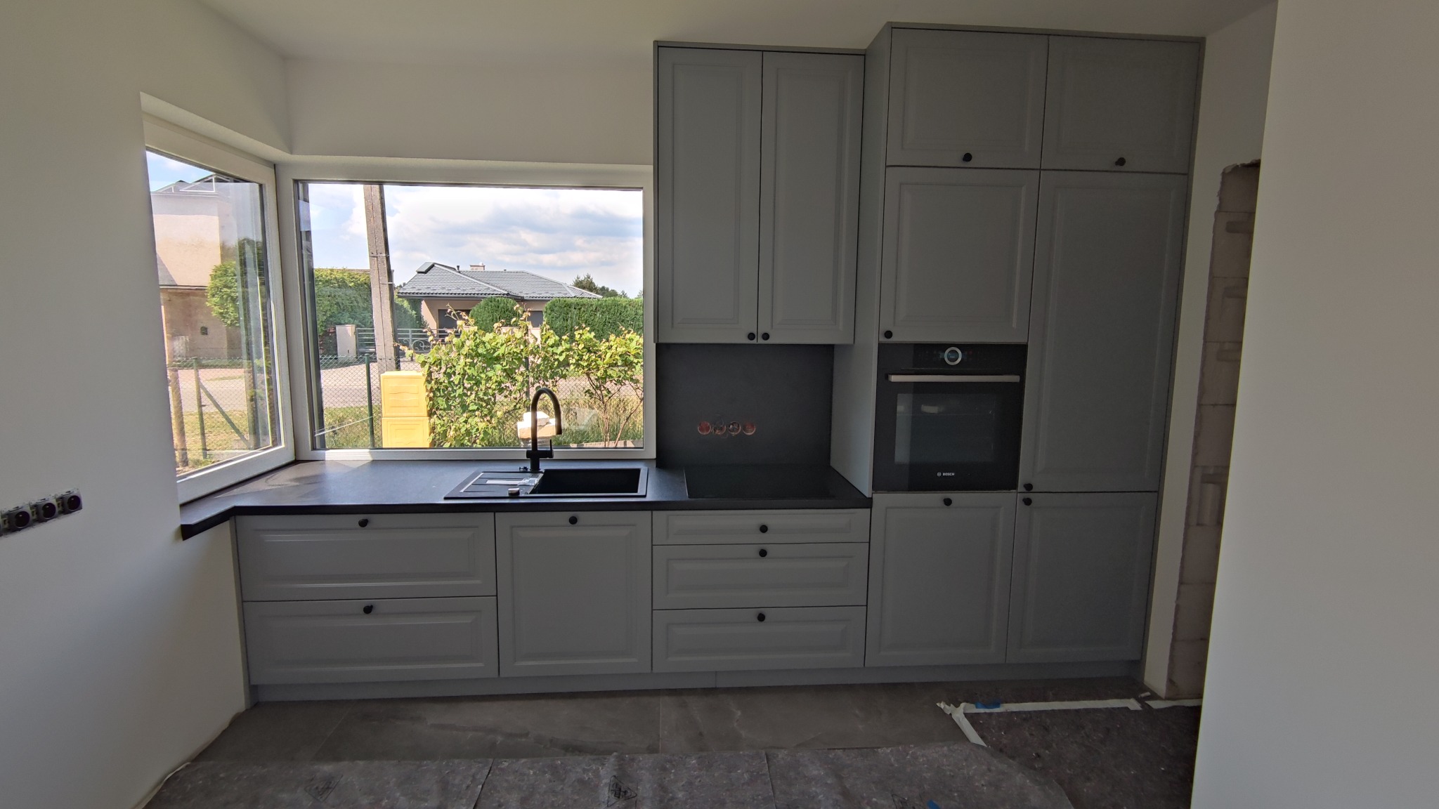 Szara zabudowa kuchenna z czarnym blatem i zlewem, widok na ogród przez okno. W tle widać zamontowany piekarnik. Ściany w trakcie wykończenia.