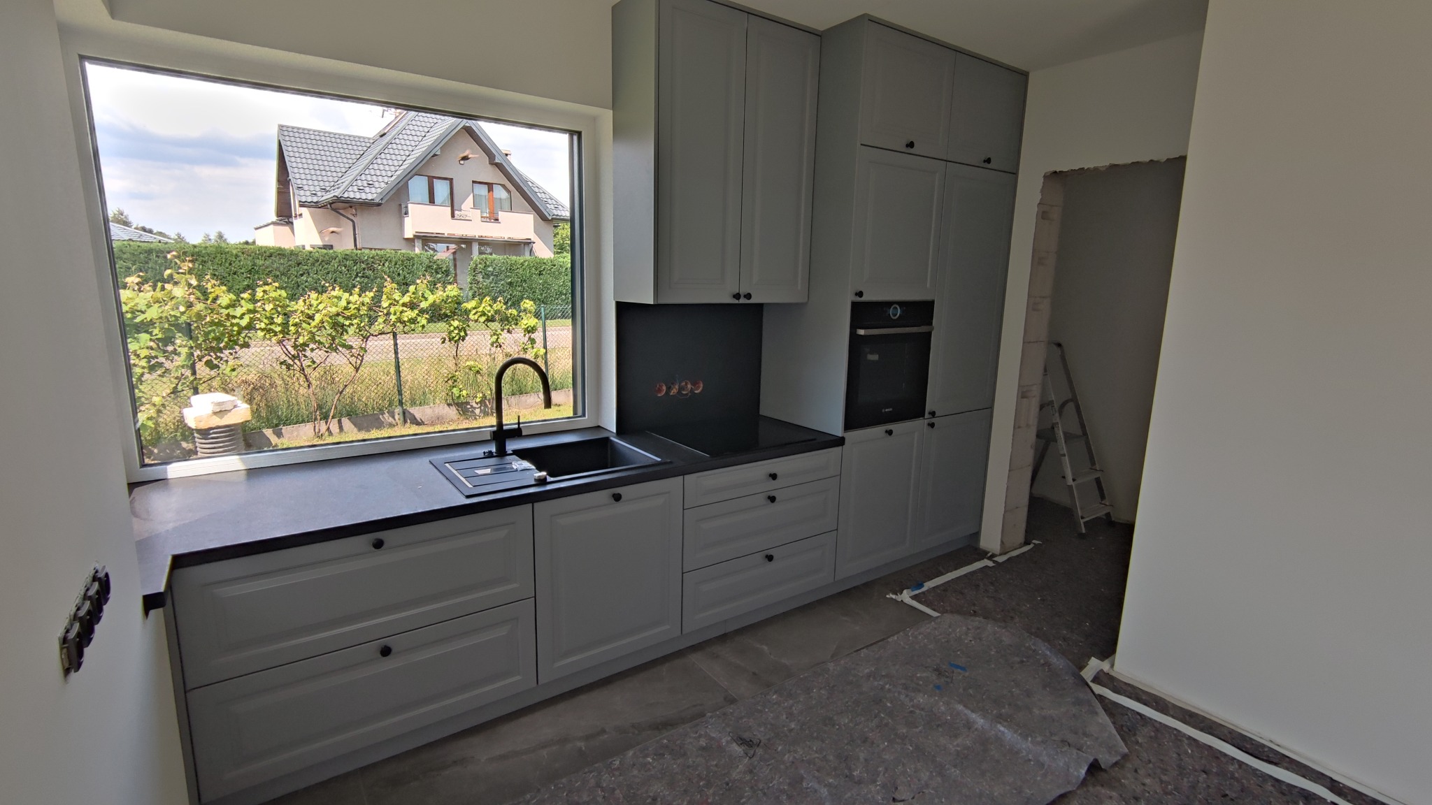 Szara zabudowa kuchenna z czarnym blatem i zlewozmywakiem pod oknem z widokiem na dom i zieleń, widoczna płyta indukcyjna i piekarnik, podłoga zabezpieczona folią malarską.