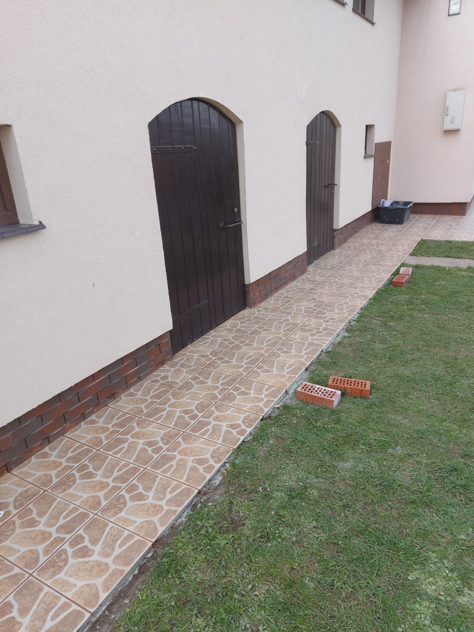 Ułożona płytkami ścieżka prowadząca do ciemnych, drewnianych drzwi w jasnej elewacji budynku, z cegłami leżącymi na trawniku obok.