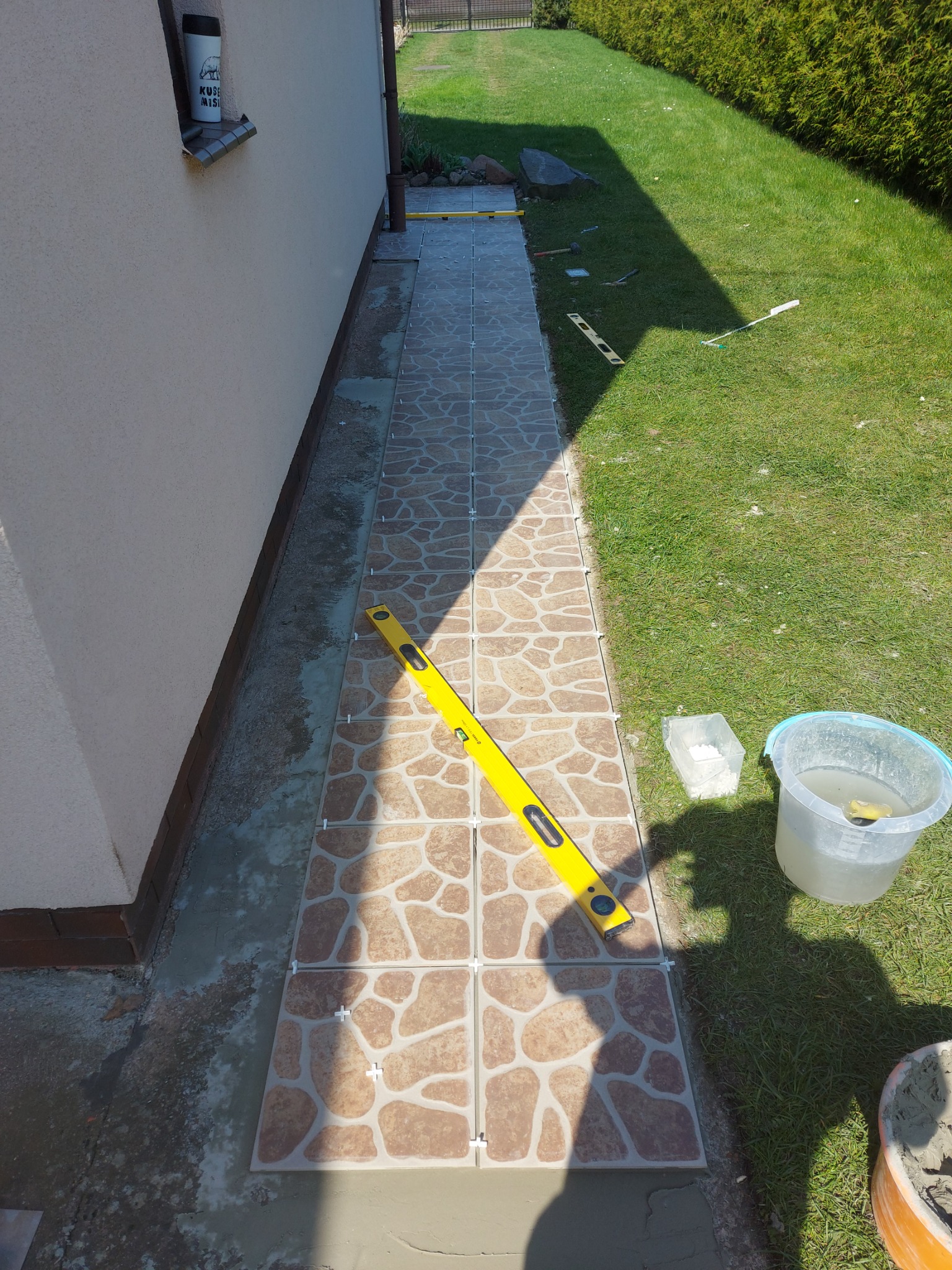 Układanie płytek na zewnątrz budynku, widoczny poziomica, wiadro z wodą i zaprawa, częściowo ułożona ścieżka z płytek.