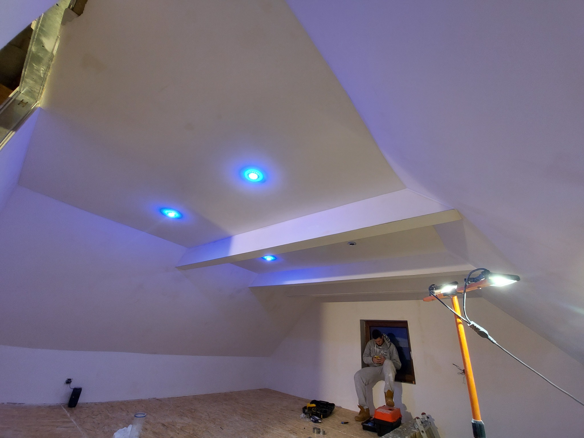 Poddasze w trakcie wykańczania: pracownik siedzi na parapecie okna, sprawdzając telefon; widoczne oświetlenie punktowe LED w suficie, ściany po szpachlowaniu, podłoga z płyt OSB oraz lampa budowlana.