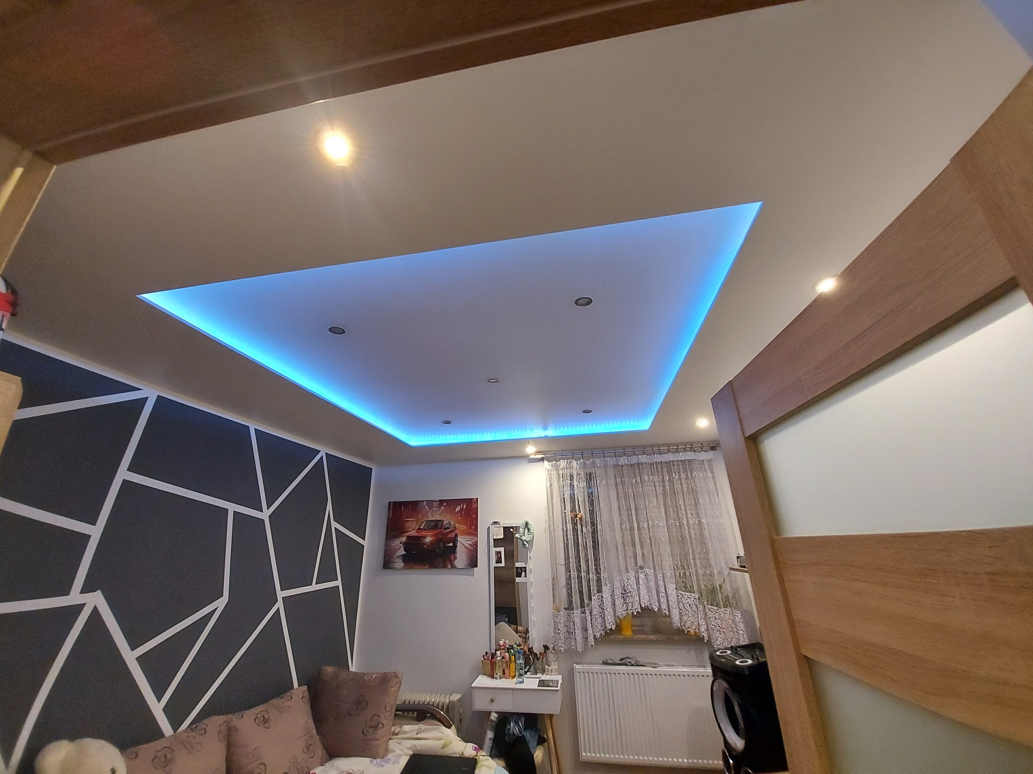 Nowoczesny sufit podwieszany z oświetleniem LED w kolorze niebieskim, widok z dołu na pokój z dekoracyjną ścianą i elementami wyposażenia.
