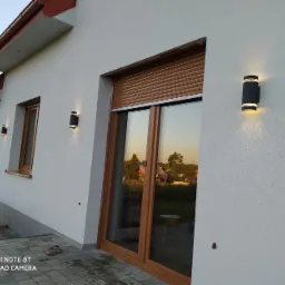 Elewacja domu z zamontowanymi czarnymi lampami zewnętrznymi świecącymi w górę i w dół, widoczne okna z brązowymi ramami i roletami zewnętrznymi, kostka brukowa na tarasie.