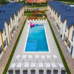 Kompleks identycznych domów z ciemnymi dachami, basenem pośrodku i rzędem leżaków, widok z góry.