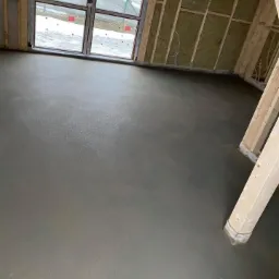 Świeżo wylana, gładka, szara posadzka w surowym wnętrzu domu w trakcie budowy, widoczne drewniane elementy konstrukcyjne ścian i okno z widokiem na zaśnieżony teren.