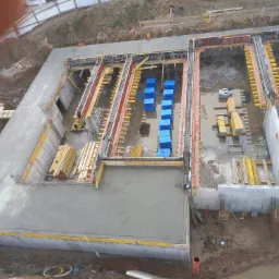 Zalewanie betonem fundamentów budynku z widocznymi szalunkami, zbrojeniami i niebieskimi opakowaniami materiałów budowlanych, widok z góry.