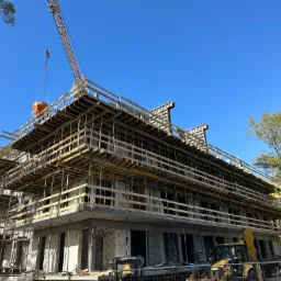 Wielopiętrowy budynek w trakcie budowy, z widocznym szalunkiem, dźwigiem podnoszącym beton oraz żółtą koparką na pierwszym planie, na tle błękitnego nieba i drzew.