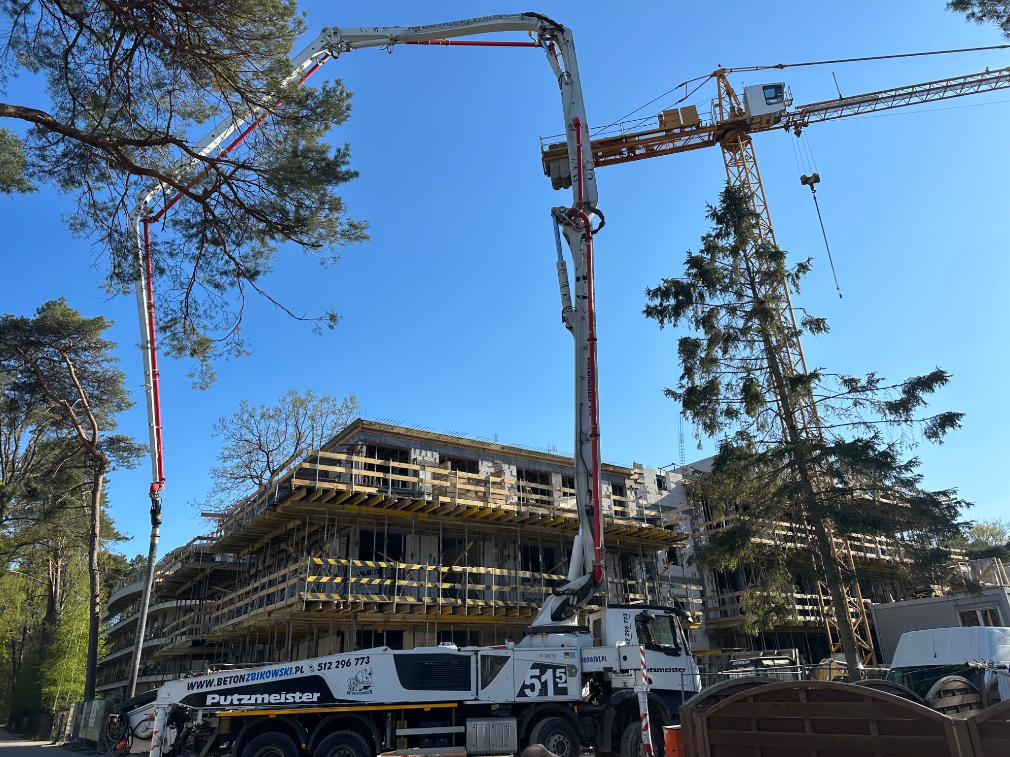 Betonomieszarka firmy Putzmeister z wysięgnikiem pompy do betonu na tle budynku w stanie surowym otwartym z żółtym szalunkiem i żurawiem budowlanym, widoczne drzewa i błękitne niebo.