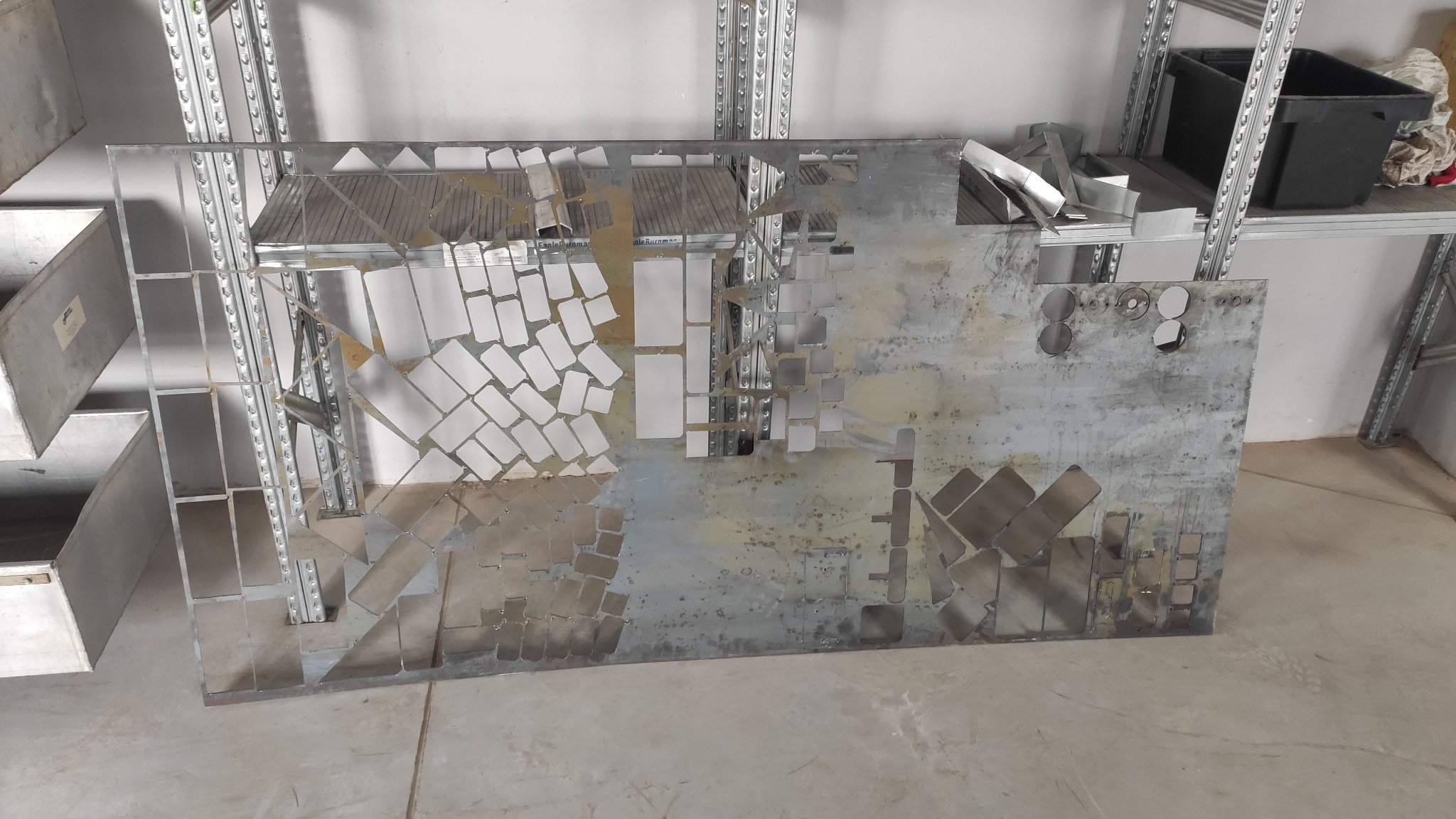 Metalowa płyta z wyciętymi wzorami geometrycznymi, leżąca w warsztacie, widoczne ślady obróbki i metalowe odpady na tle regałów.