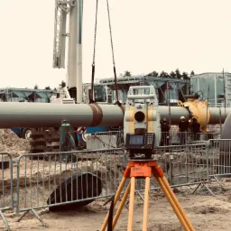 Pomarańczowy teodolit z ekranem na trójnogu, na pierwszym planie wykop z metalowym ogrodzeniem, w tle rurociąg podnoszony przez dźwig i pracownicy w kaskach.