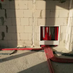 Świeżo ułożone, czerwone rury grzewcze wychodzące ze ściany z bloczków betonowych, zabezpieczone pianką montażową, widoczne również rury karbowane w podłodze.